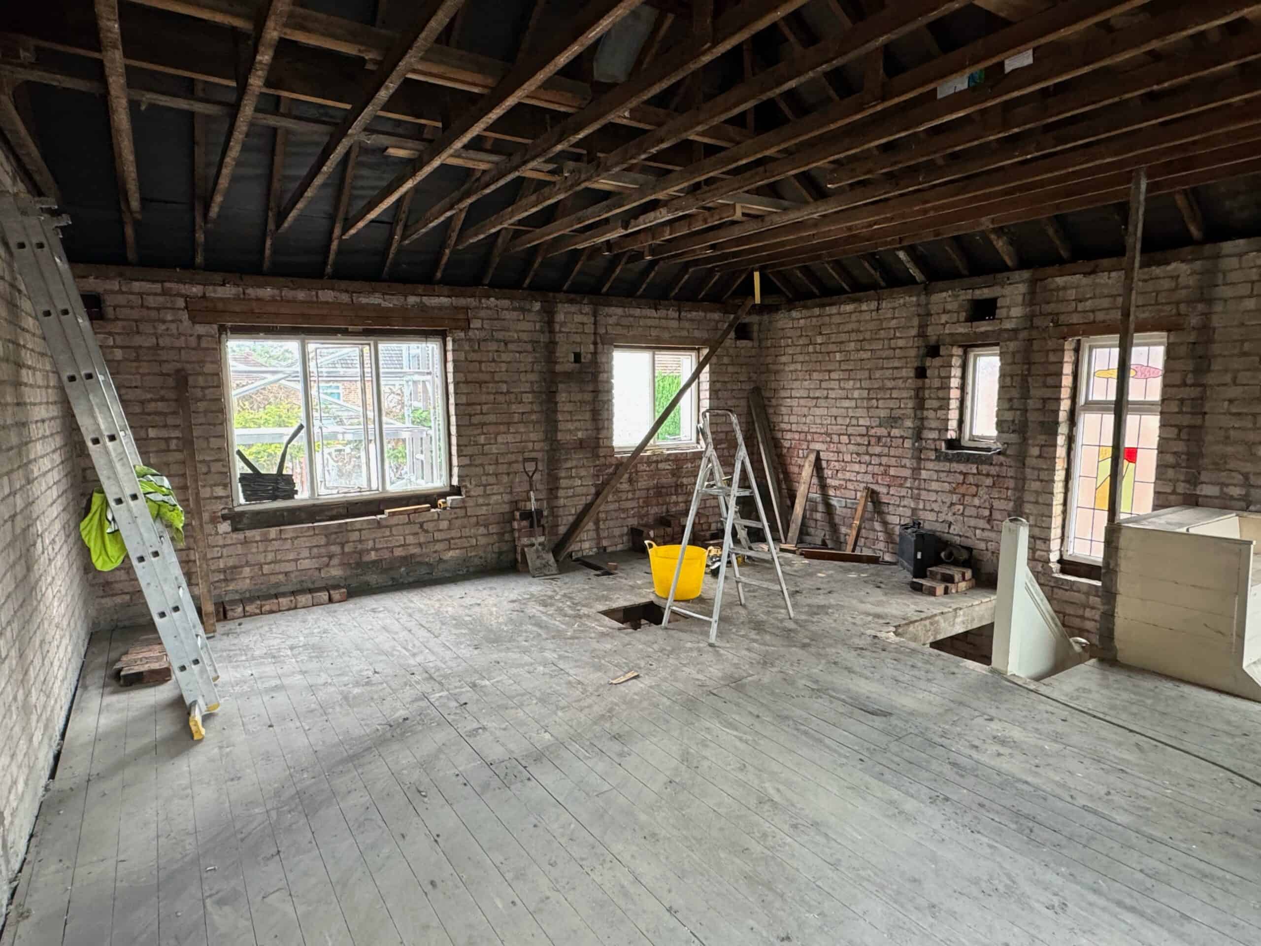 An empty room under renovation with exposed brick walls and wooden ceiling beams. Two ladders and a yellow bucket are in the center, and there are upper-level windows letting in natural light. Floorboards are visible, and various tools are scattered around.