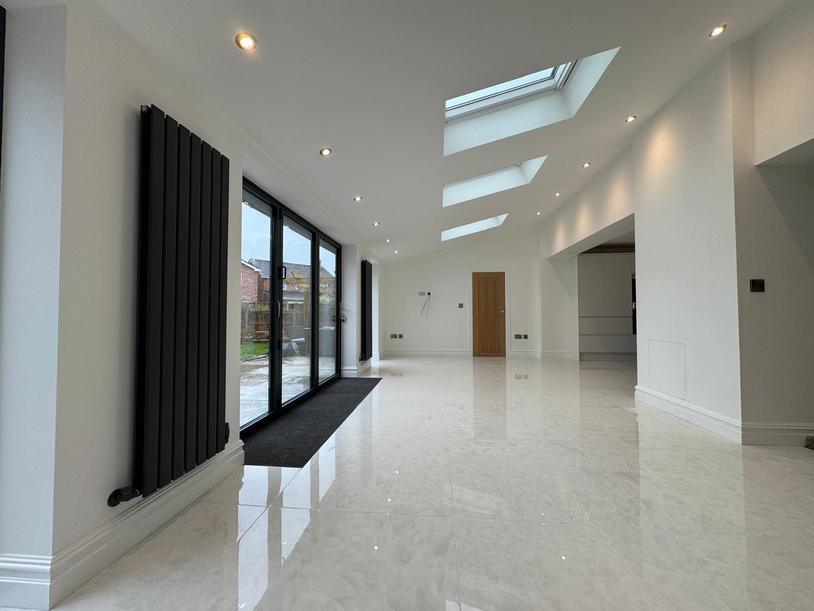 A spacious, modern room with glossy white tiled floors, white walls, and several skylights. Large glass doors open to an outdoor area. A vertical radiator is mounted on the wall beside the doors.