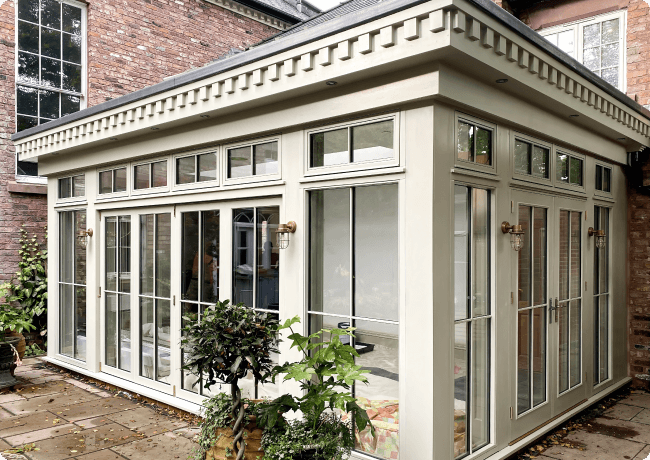 A sunroom with large glass windows and a flat roof is attached to a brick house. Potted plants surround the patio area, and stylish outdoor lanterns are mounted on the sunroom walls.