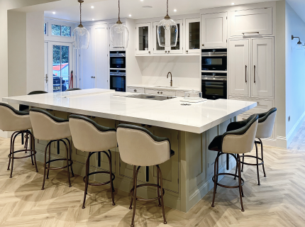 Modern kitchen with a large white island, surrounded by six beige bar stools. The island features a stovetop and sink. White cabinets, ovens, and pendant lights are visible. The floor has a herringbone pattern.