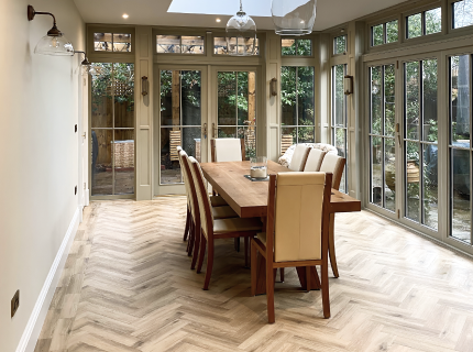 A bright dining room featuring a wooden table with eight cream-colored chairs. The room has large panel windows and doors opening to a garden view, with herringbone-patterned wooden flooring and pendant lights.