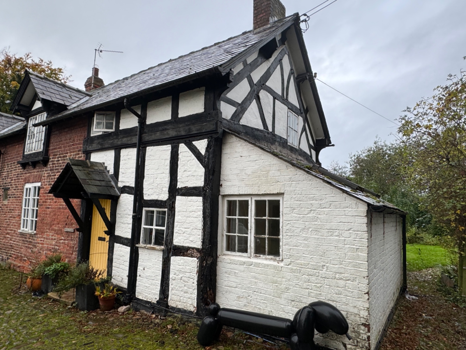 A quaint house with brick and white-painted timber framing. It has a yellow door and a small sloped roof at the entrance. A black statue resembling a large dog is on the cobblestone path. Trees and overcast skies are in the background.