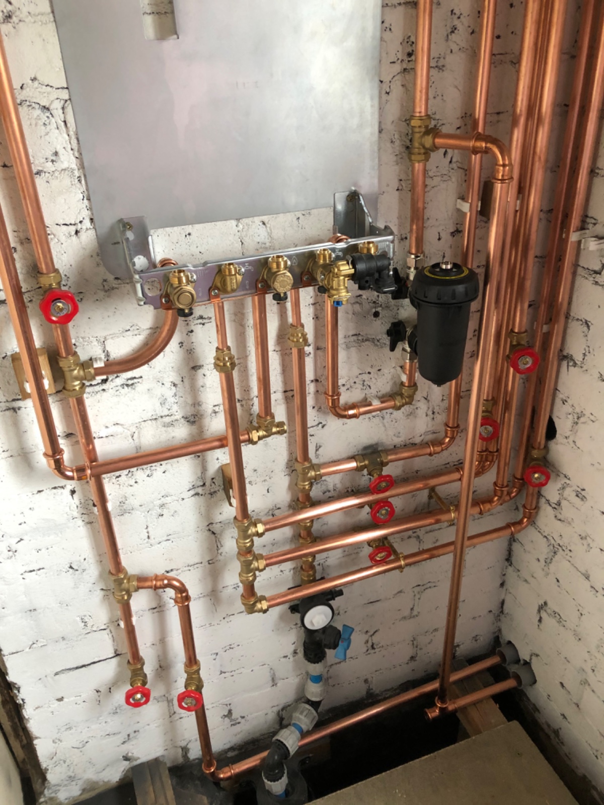 A setup of neatly arranged copper plumbing pipes with red valve handles against a white brick wall. A black filter attachment is visible, and various fittings and connections are present.