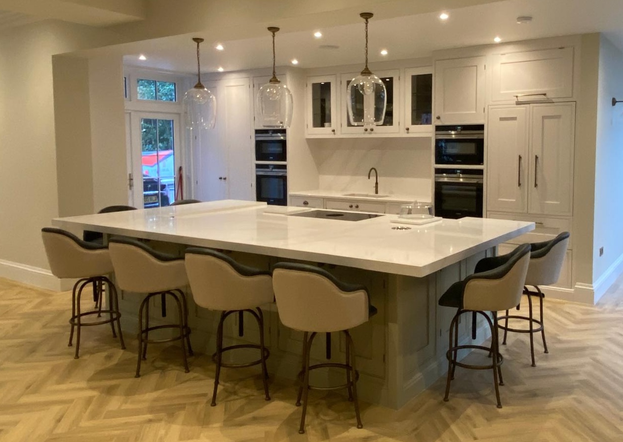 A spacious kitchen with a large central island featuring a white countertop and six cushioned stools. Behind the island are white cabinets, a sink, ovens, and pendant lights. The floor has a light wood pattern, and there's a door leading outside.