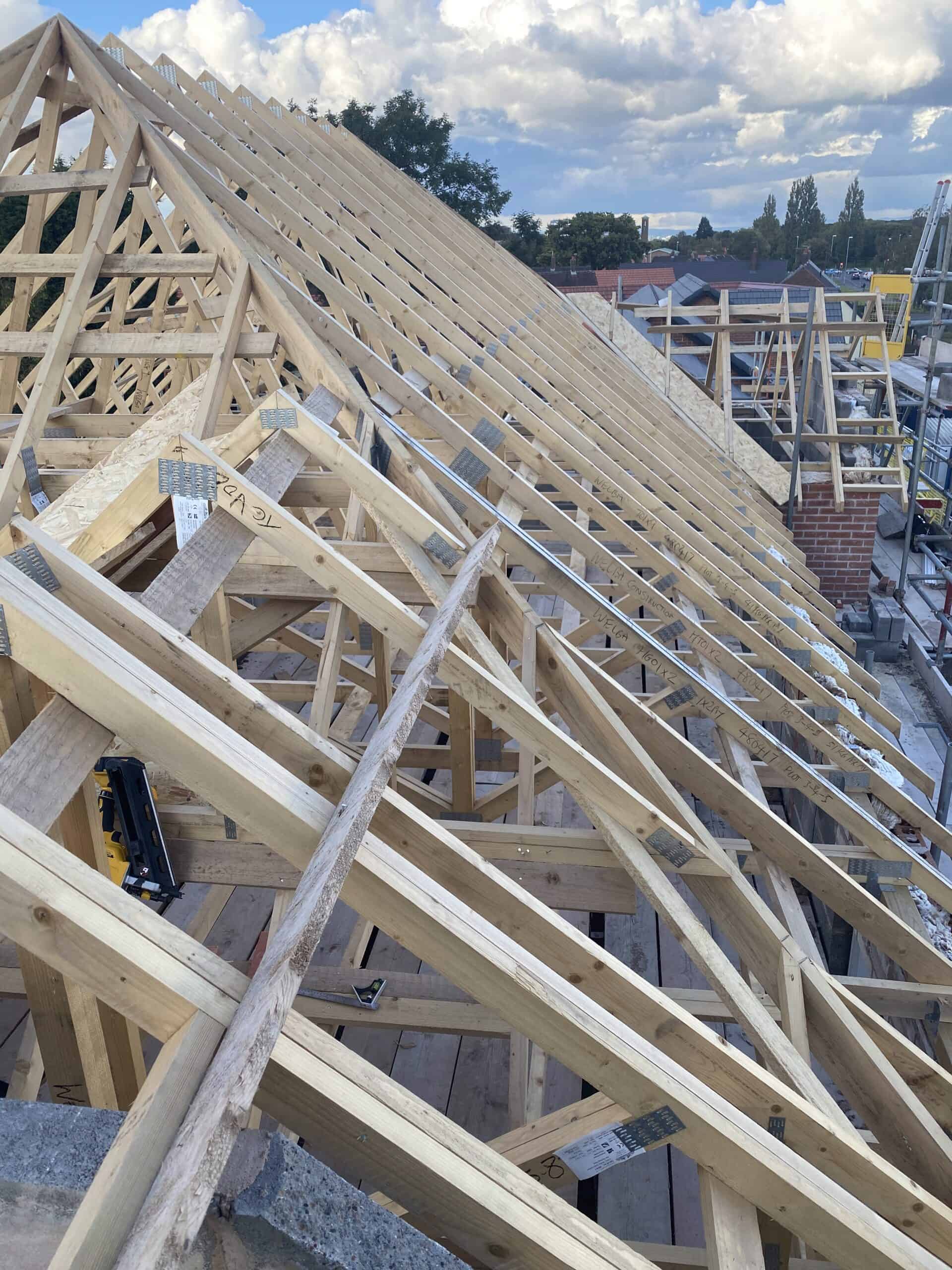 Wooden roof trusses under construction at a building site, with a clear sky and clouds in the background. The framework shows a complex pattern of beams and supports, with some brickwork visible on the side.