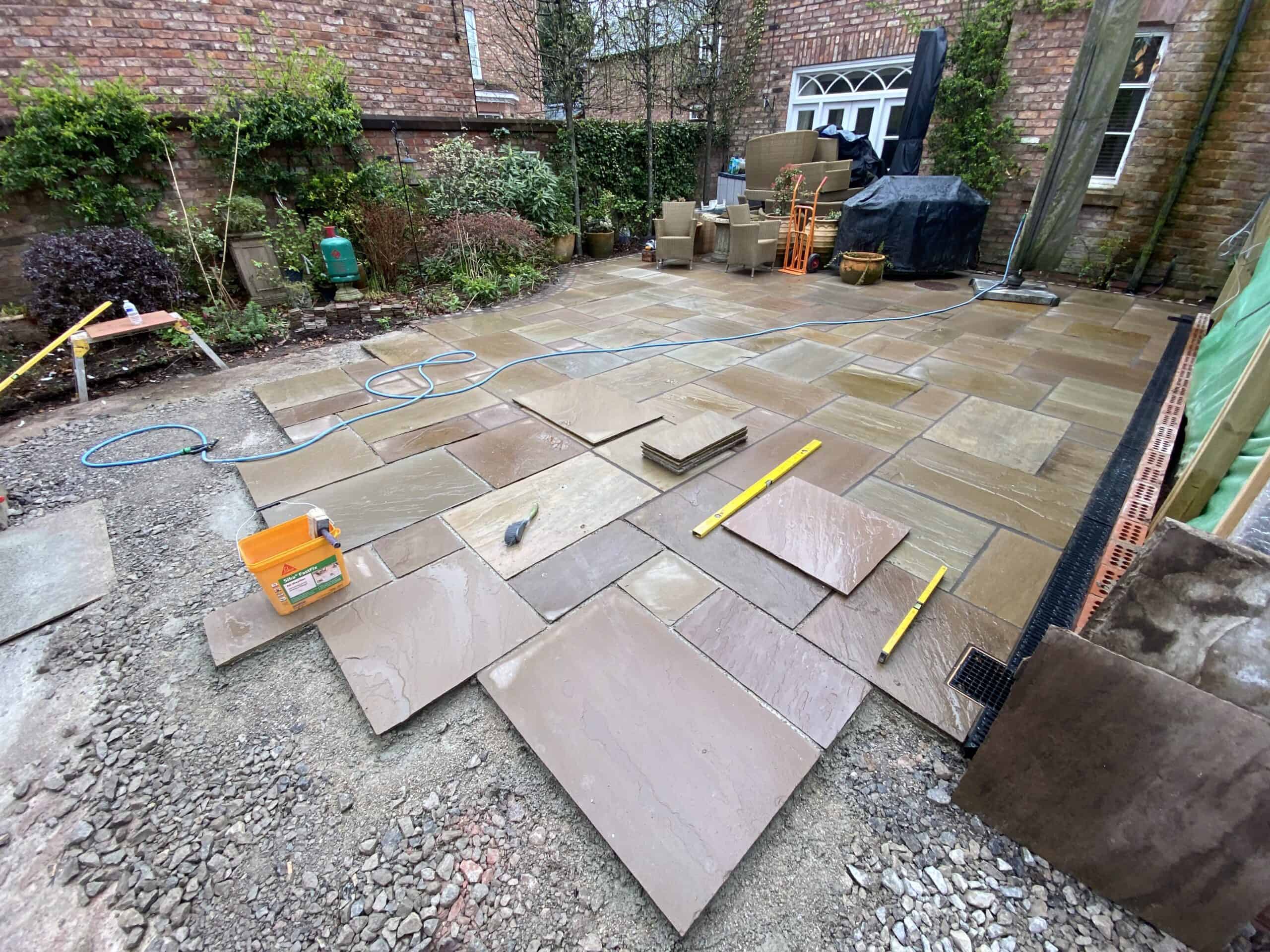 A garden patio under construction with large stone tiles being laid on the ground. Various tools like a spirit level, a hammer, and a saw are scattered around. In the background, there are stacks of materials and a partially finished area.