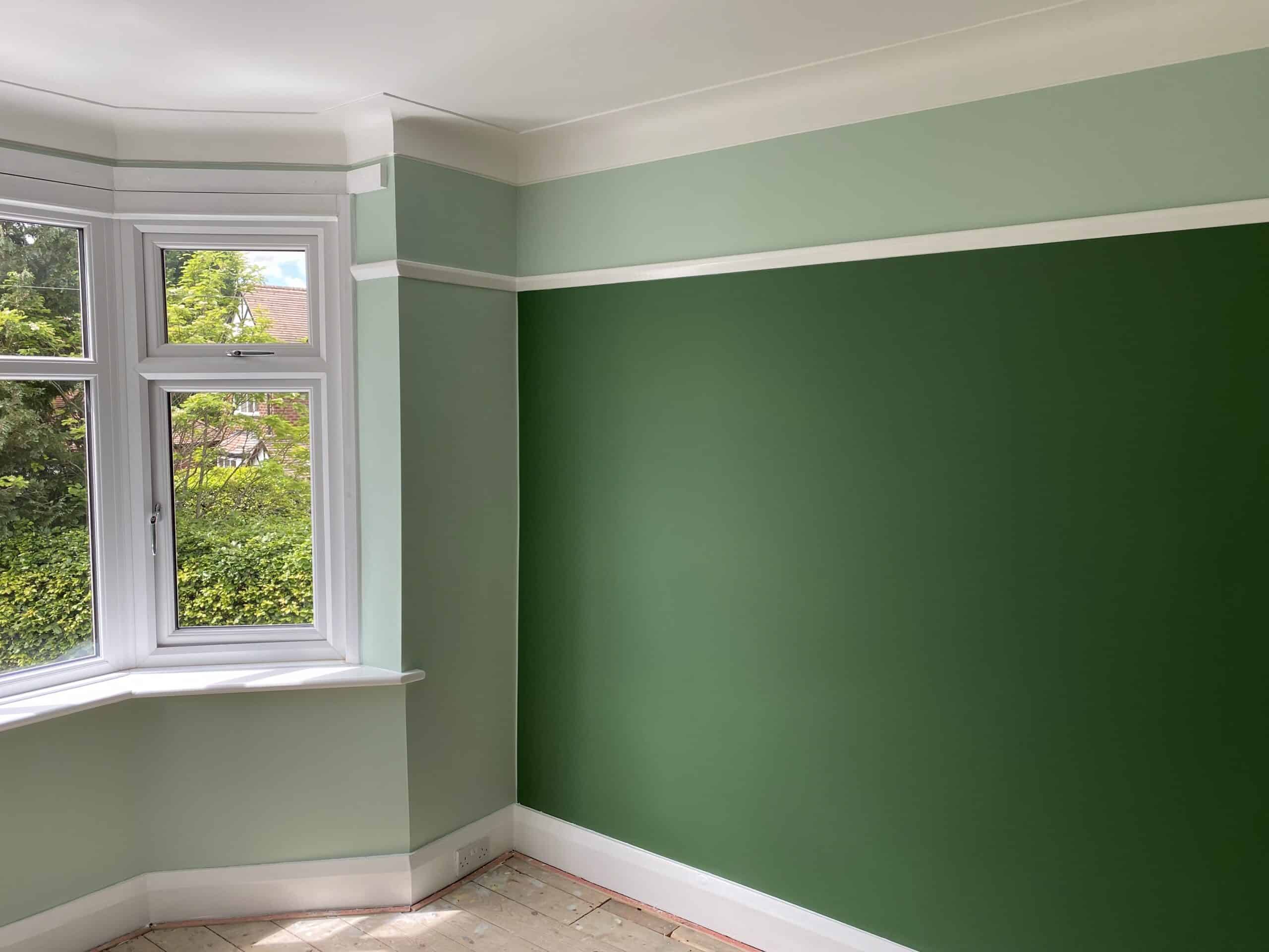 A room with freshly painted walls, featuring a large window. The walls are two shades of green: a lighter green on three walls and a darker green accent wall. White trim highlights the window and wall edges. The floor is unfinished.