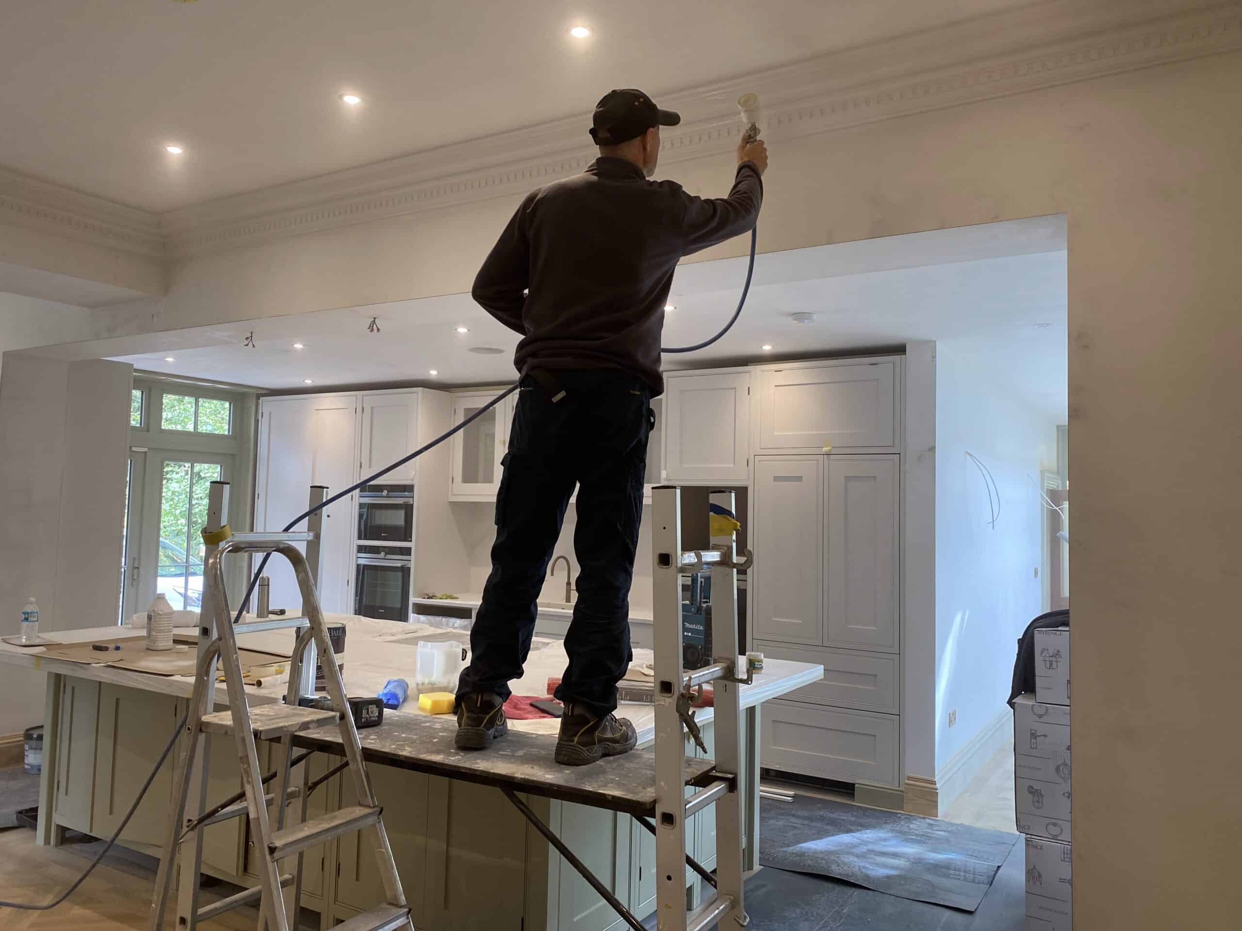 A person stands on a ladder in a kitchen, using a paint spray gun to paint the ceiling. The kitchen features a large island and built-in cabinets. Various tools and materials are scattered around, indicating ongoing renovation work.