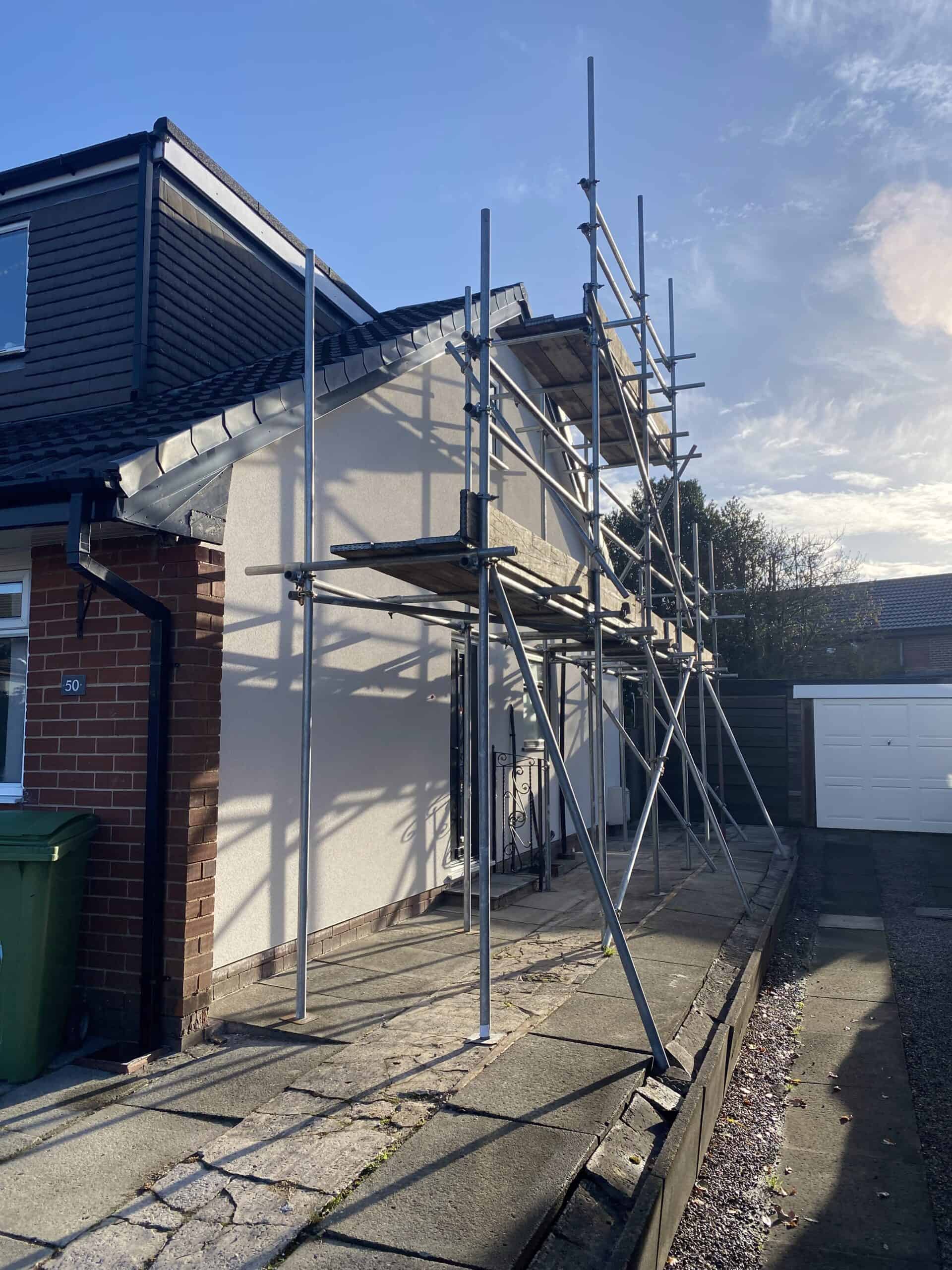 Scaffolding set up against the side of a two-story house, with a white exterior and brick accents. The scaffolding is positioned on a concrete path, extending up to the roof. A garage and trash bin are visible in the background, under a partly cloudy sky.